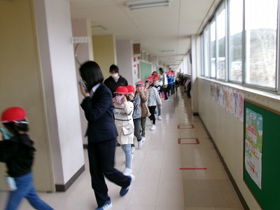 小学校校舎の廊下にて、赤い帽子を被った児童たちと、黒いスーツを着た女性教諭が避難訓練を行なっている様子を写した写真