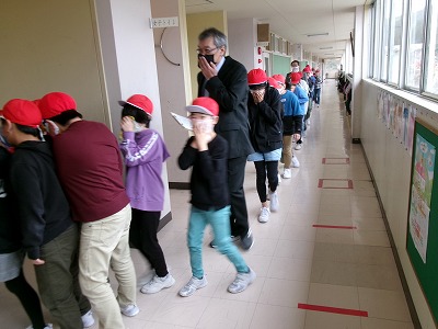 小学校校舎の廊下にて、赤い帽子を被った児童たちと、黒いスーツを着た壮年の男性教諭が避難訓練を行なっている様子を写した写真