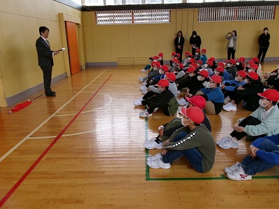 小学校の体育館にて、避難訓練を終えた赤い帽子を被った大勢の児童達が床に座りながら、黒いスーツを着た男性の話を聞いている様子を写した写真
