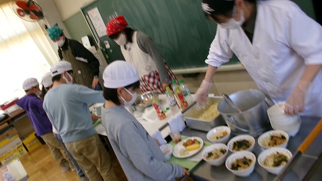 並べられた多くの給食から自分が食べられる分を選ぶ児童たちと、それを手伝う3名の先生たちの写真