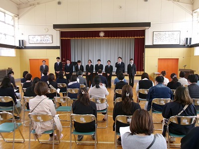 小学校の体育館で行われたPTA総会にて、整然と並べられたパイプ椅子に座る大勢の保護者の人達と、壇上の前に立つスーツ姿の男性9人の様子を写した写真