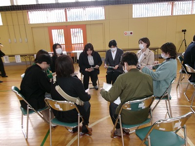 小学校の体育館で行われたPTA総会にて、小さな円形に並べられたパイプ椅子に座って会議を行う、9人の保護者の様子を写した写真