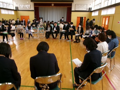 小学校の体育館で行われたPTA総会にて、部会ごとに円形に並べられたパイプ椅子に座って、手元の資料を見ながら会議を行う、大勢の保護者の様子を写した写真