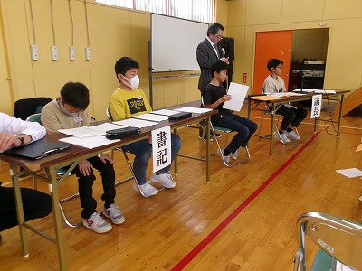 小学校の体育館で行われた児童会総会にて、「書記」の席に座る2人の男子児童と、その隣に座る1人の男子児童、その右奥の「議長」の席に座る1人の男子児童の様子を写した写真