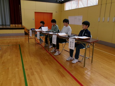 小学校の体育館で行われた児童会総会にて、役割に応じて決められた席に座る4人の男子児童の様子を写した写真