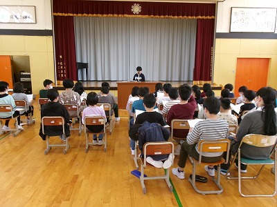 小学校の体育館で行われた児童会総会にて、整然と並べられた学校椅子に座る大勢の児童達と、彼らと向かい合って演壇で話をする1人の児童の様子を写した写真