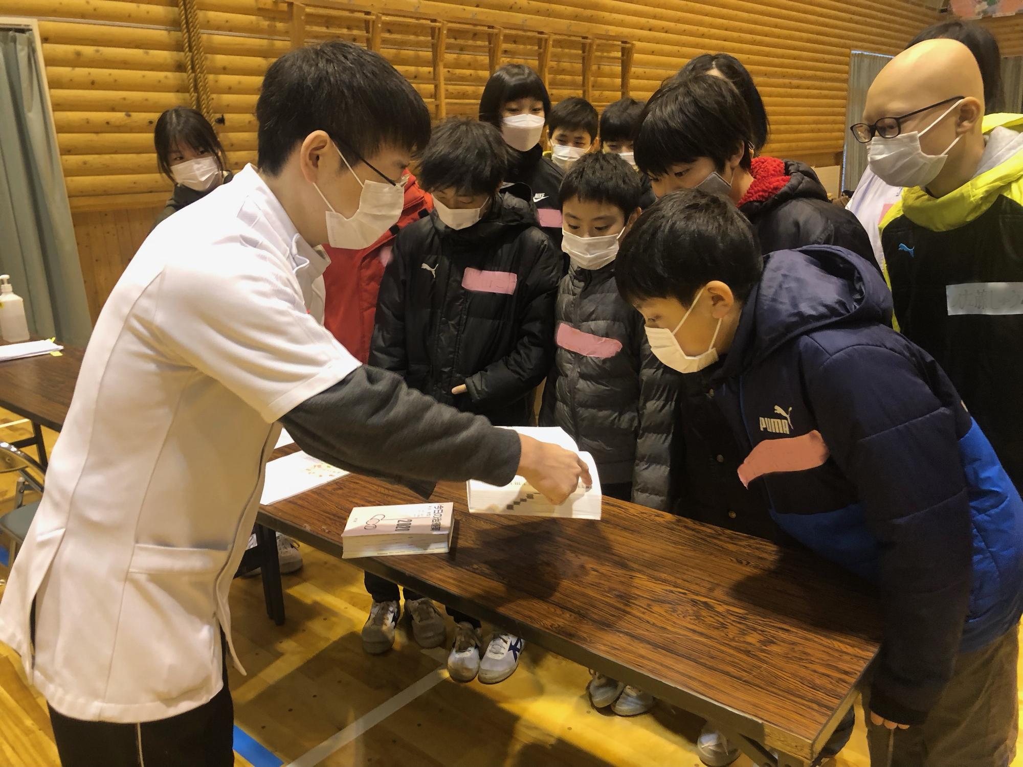 本を見せながら薬剤師の仕事について説明する男性と本を興味深くのぞき込む生徒たちの写真