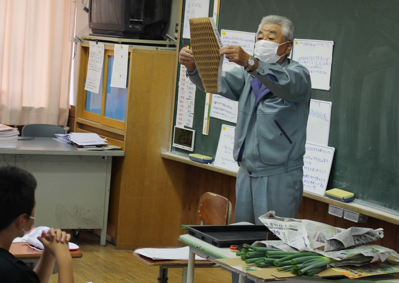 学校の教室で、作業服を着た男性が包装用の紙製ネットを広げている写真