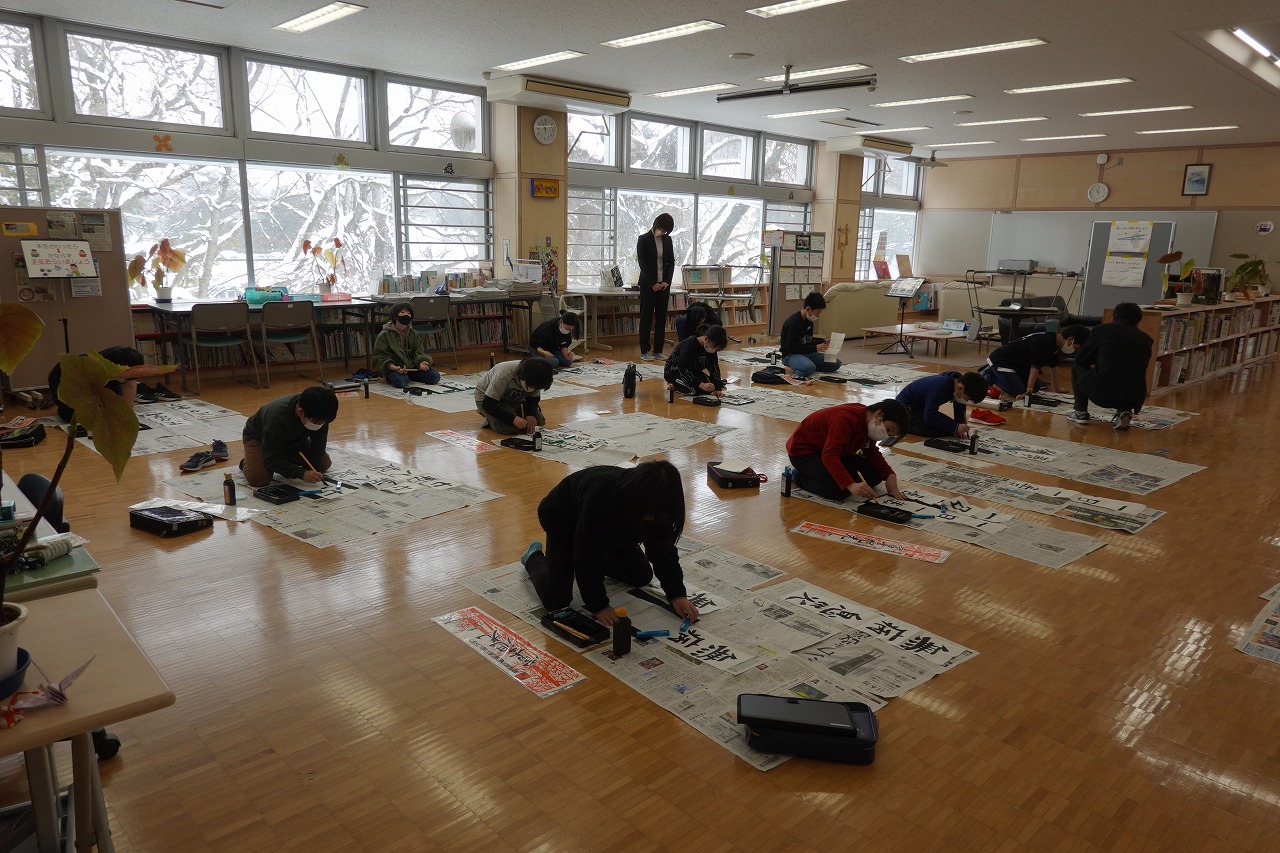 広い会場で、床に新聞紙を敷いて書き初め会を行う生徒たちの写真