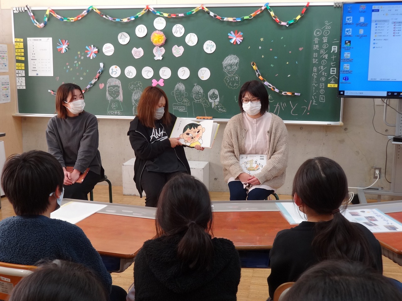 小学校の教室内で、黒板の前で座って絵本の読み聞かせを行うPTA研修部の3人の母親と、向かい合って座りながら話を聞く、複数人の児童達の様子を写した写真
