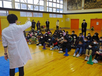 体育館に集まった児童たちの前で科学の授業をする、最上広域市町村圏事務組合教育研究センターの男性の先生の写真