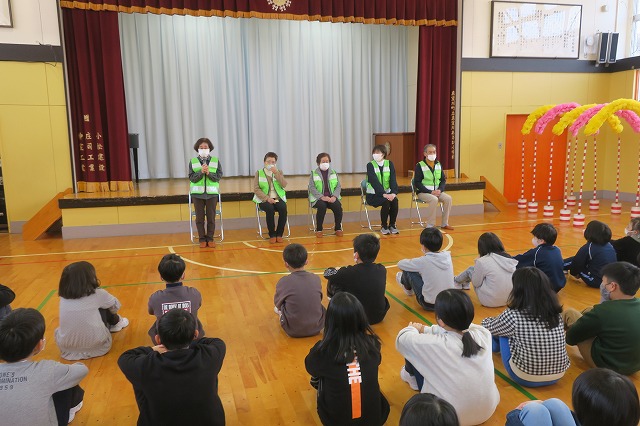 そろいのライトグリーンのベストを着た5人の「見守り隊」の皆さんと、体育館でそれを迎える児童たちの写真