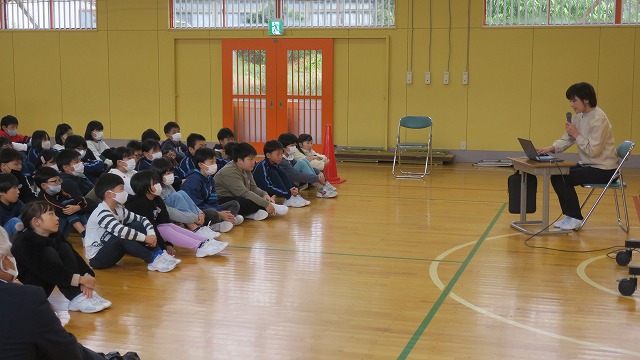 体育館で先生が栄養バランスの大切さについて説明し、それを座って熱心に聞いている生徒たちの写真