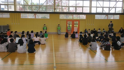 小学校の体育館で実施された見守り隊の顔合わせ会にて、体育館の床に座る大勢の児童達と、向かい合って立つ3人の見守りたいメンバーを写した写真