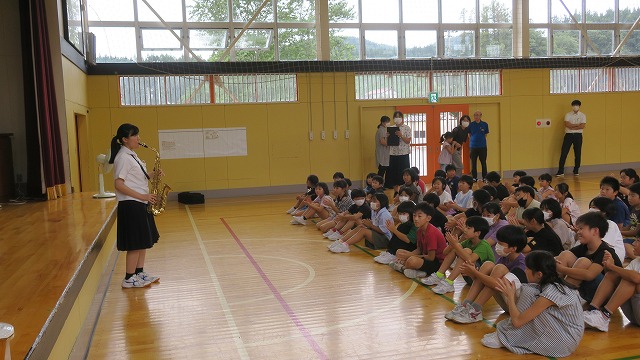 体育館でサックスの演奏を披露する女子中学生と熱心に聴く児童たちの写真