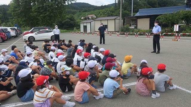 学校の駐車場で前に立って地震を想定した避難訓練について説明する先生とそれを座って聞く生徒たちの写真