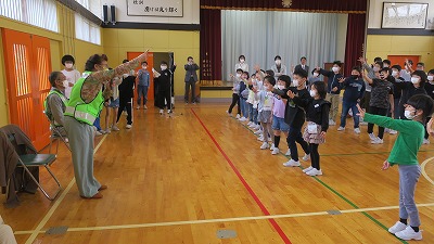 小学校の体育館で実施された見守り隊の顔合わせ会にて、見守り隊のメンバーの男性が、大勢の児童達と向かい合ってじゃんけんをする様子を写した写真