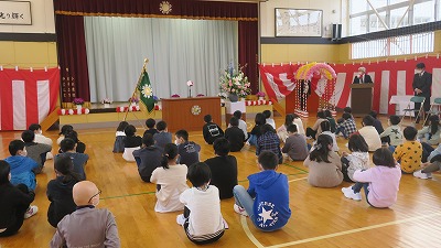 体育館で演台から話す校長先生と床に体育座りをして話を聞く児童たちの写真