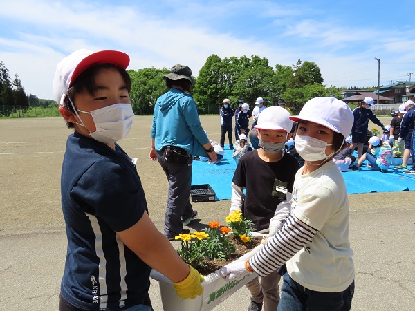 赤白帽子を被った子供が3人で花が植えられたプランターを運んでいる写真