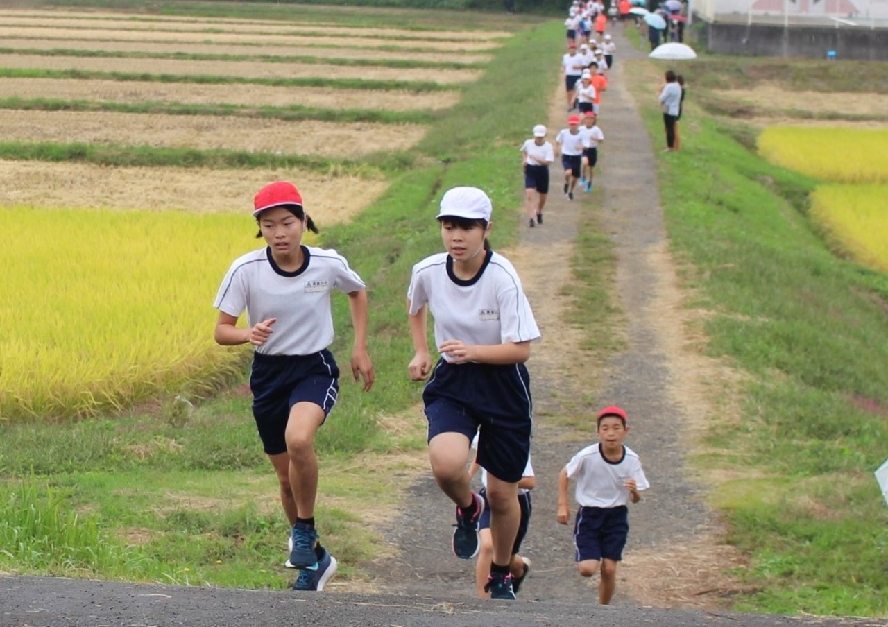田んぼの間の坂道を体操服姿の子供たちが走っている写真