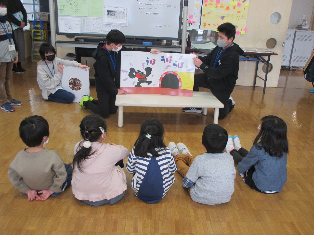 小学校の教室内で行われた新入生体験入学にて、座っている5人の入学前の児童の前で、絵本を使って読み聞かせを行う5年生の3人の児童の様子を写した写真