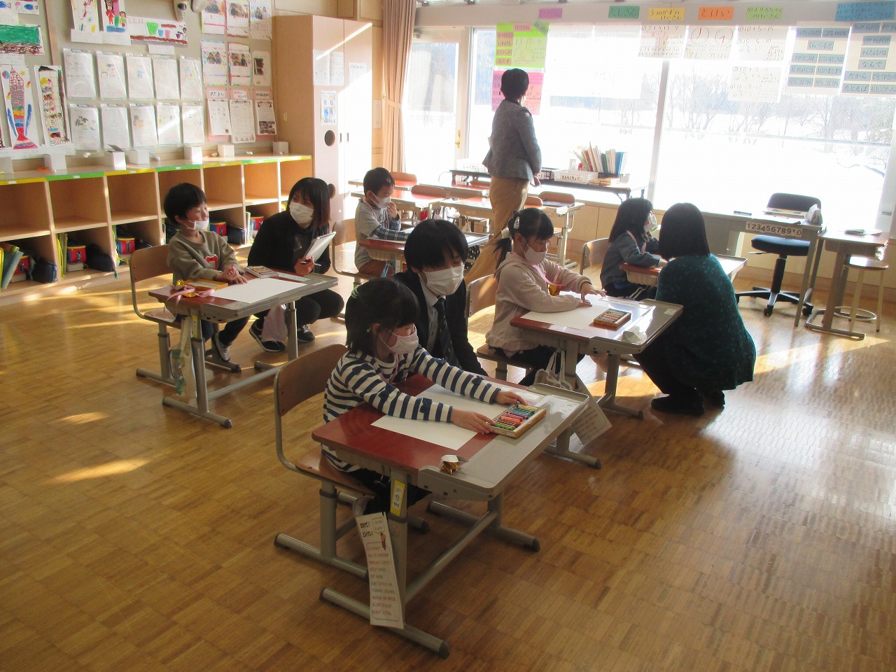 小学校の教室内で行われた新入生体験入学にて、教室の椅子に座って、4人の教職員から絵の描き方を学んでいる5人の入学前の児童の様子を写した写真