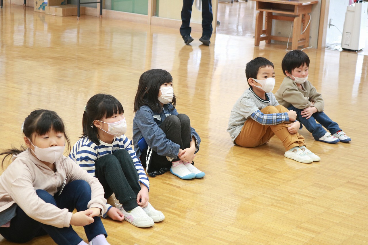 小学校の教室内で行われた新入生体験入学にて、教室の床に体育座りをしている5人の入学前の児童の様子を写した写真