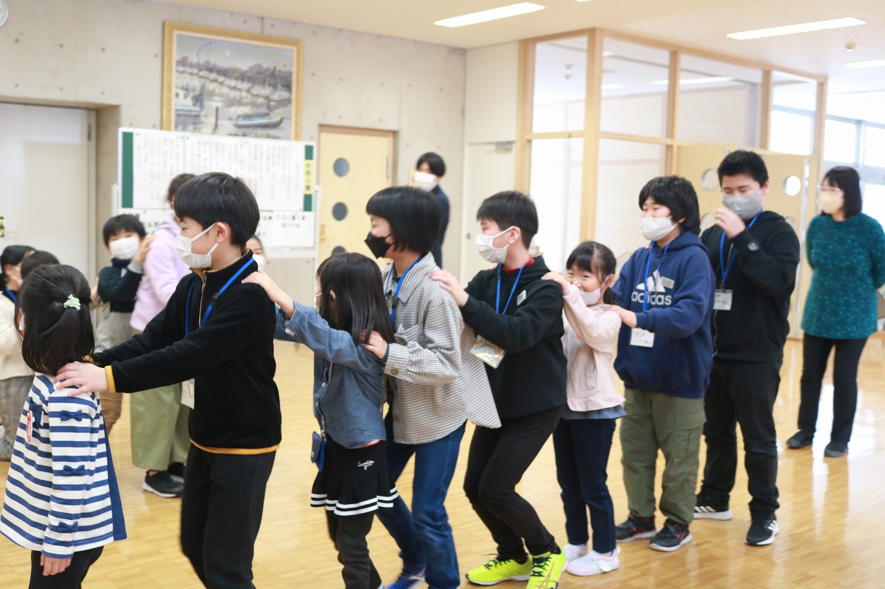 小学校の教室内で行われた新入生体験入学にて、5年生と入学前の児童達が、各々前の児童の肩を持ちながら隊列を組んで遊んでいる様子を写した写真