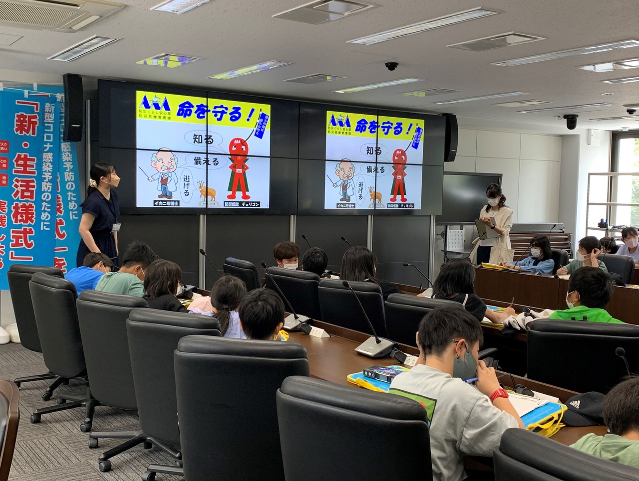 「命を守る!」という文字等が映ったスクリーンの横で説明している女性と席に着いてそれを聞いている子供たちの写真