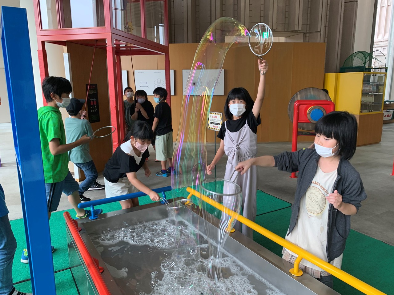 子供たちがしゃぼん液の入った水槽に鉄で作った輪を通して大きなシャボン玉を作っている写真