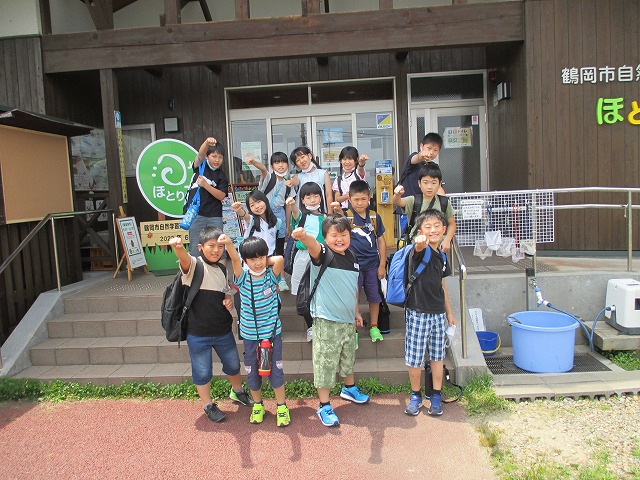 校外学習にて、鶴岡市自然学習交流館「ほとりあ」の入口前の階段で、3年生の児童13人が、階段の上から3列に並んで、腕を前に突き出すポーズを取っているところを撮影した記念写真