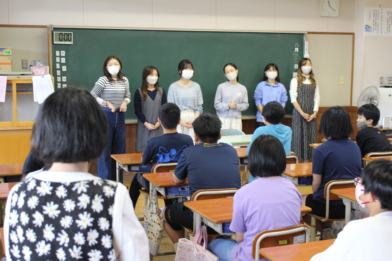 黒板の前で6人並んだ女性を席についた子供たちが眺めている写真