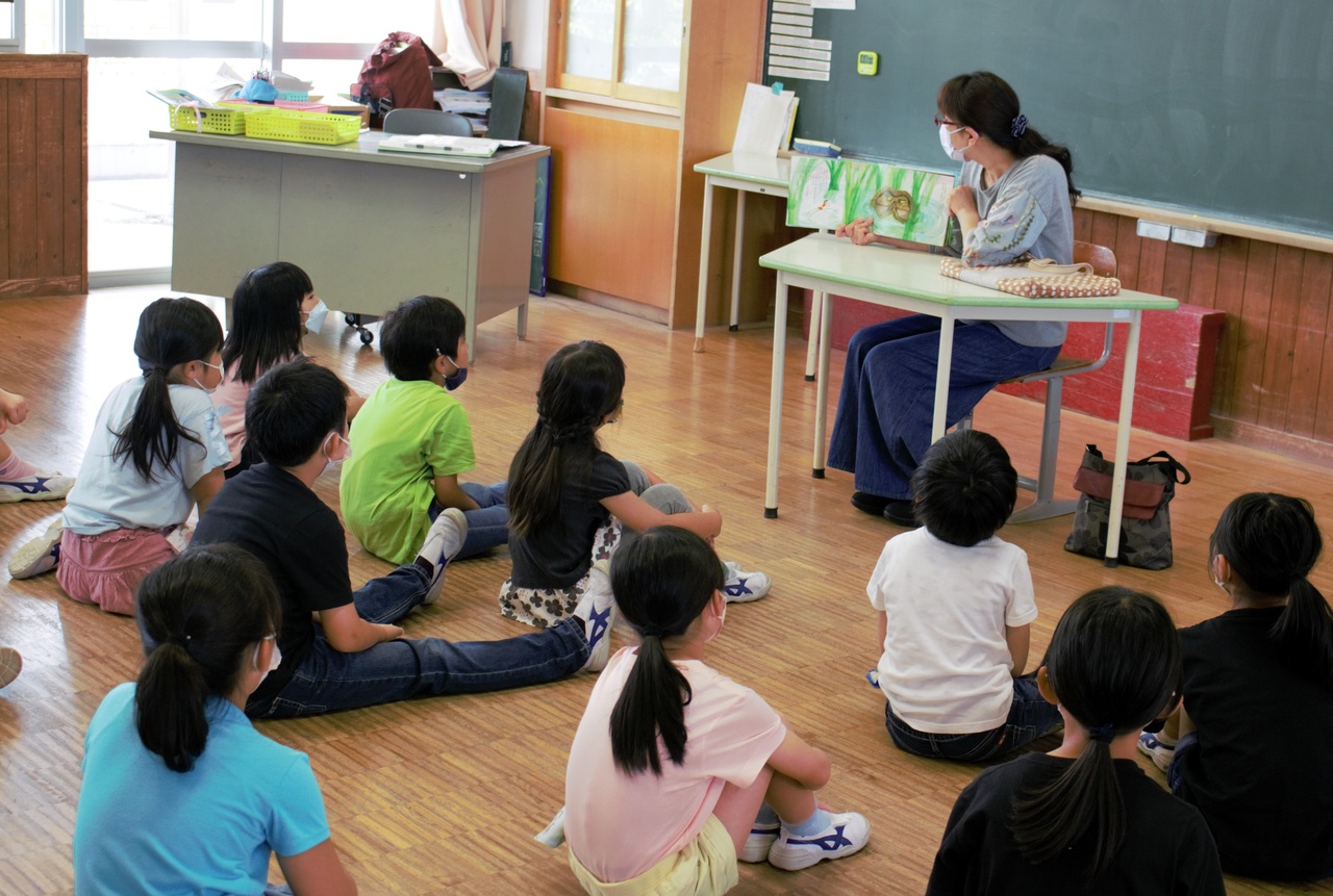 床に座った子供たちが机の上に絵本を乗せて読み聞かせをしている女性を眺めている写真