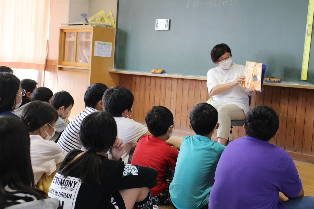 床に座った子供たちが椅子に座って絵本の読み聞かせをしている女性を眺めている写真