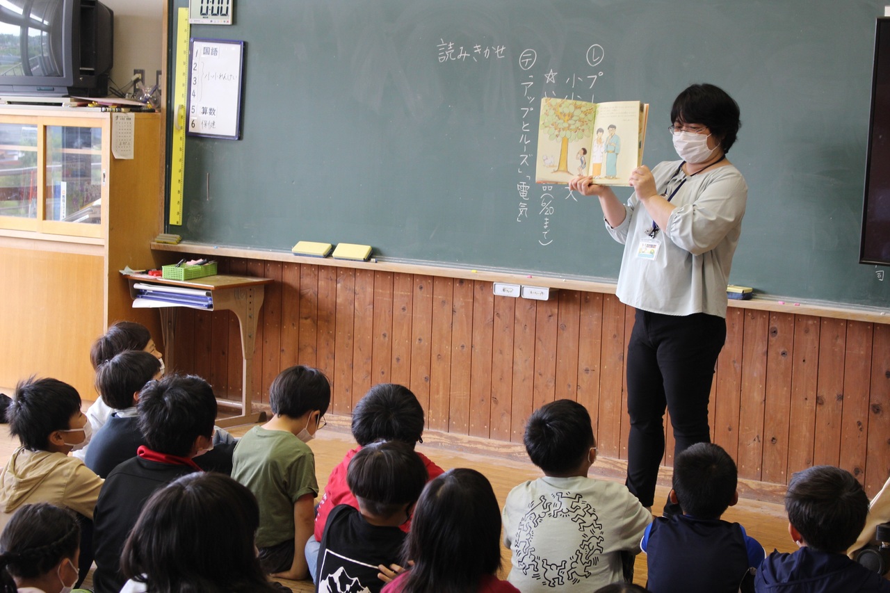 床に座った子供たちが立って絵本の読み聞かせをしている女性を眺めている写真