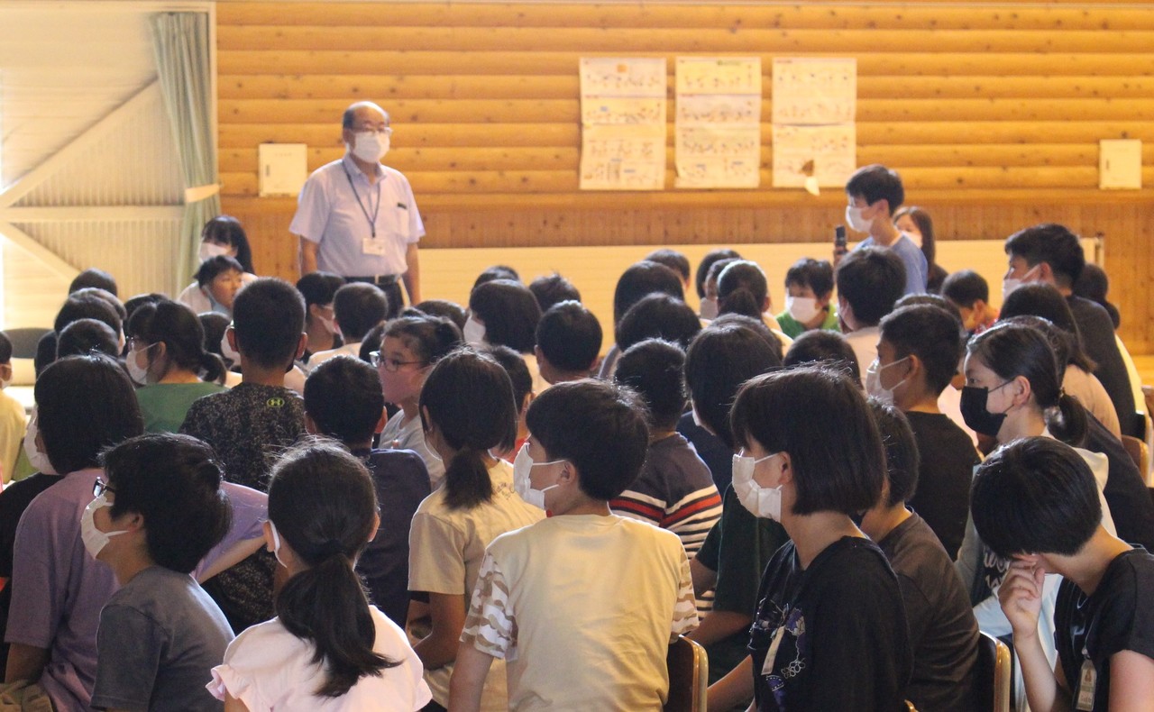 体育館で椅子に座った大勢の子供たちの中で1人立っている男の子に、首からカードを下げた男性が目を向けている写真