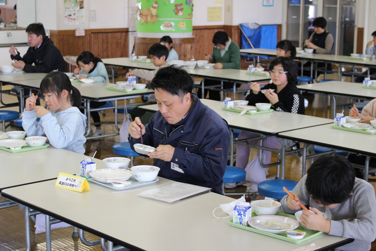 机につき給食を食べる男性と子供たちの写真