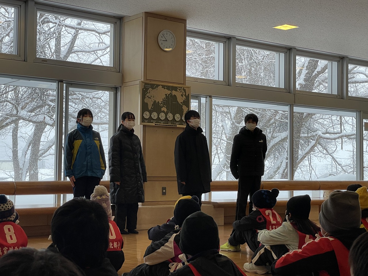 真室川北部小学校スキー大会にて、スキー場施設内で、床に座る大勢の児童達から見送りを受ける、スキー大会参加者の高学年の児童4人の様子を写した写真