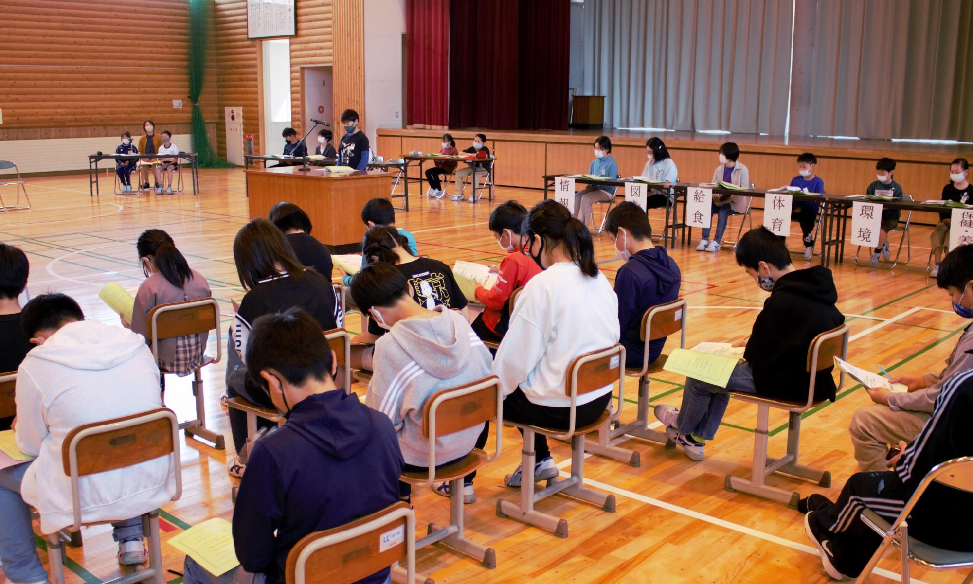 体育館に置かれた講演台で子供が話しているのを手元のプリントを見ながら聞く子供たちの写真