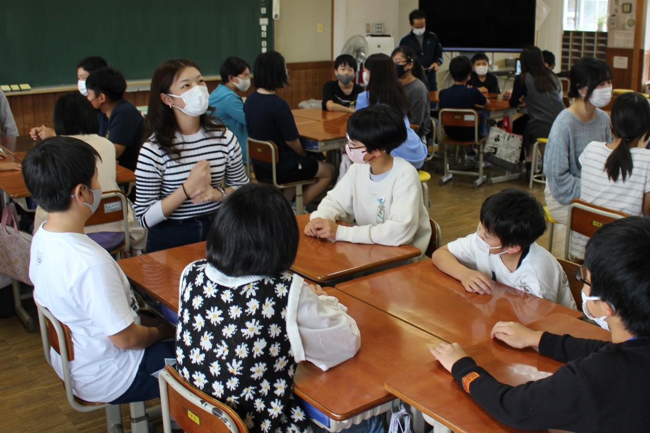 寄せられた机についた5人の子供が女性の話を聞いている写真