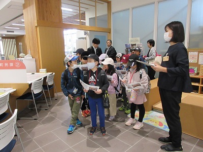 役所の中で子供たちが、胸に名札を付け書類を持った女性の説明を聞いている写真