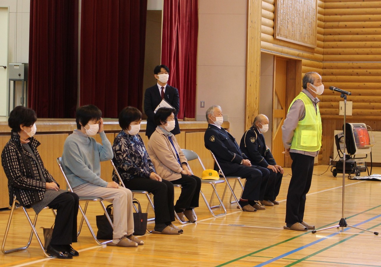 体育館の前方でパイプ椅子に座っている人たちとマイクの前で話している黄色のベストを着た男性の写真