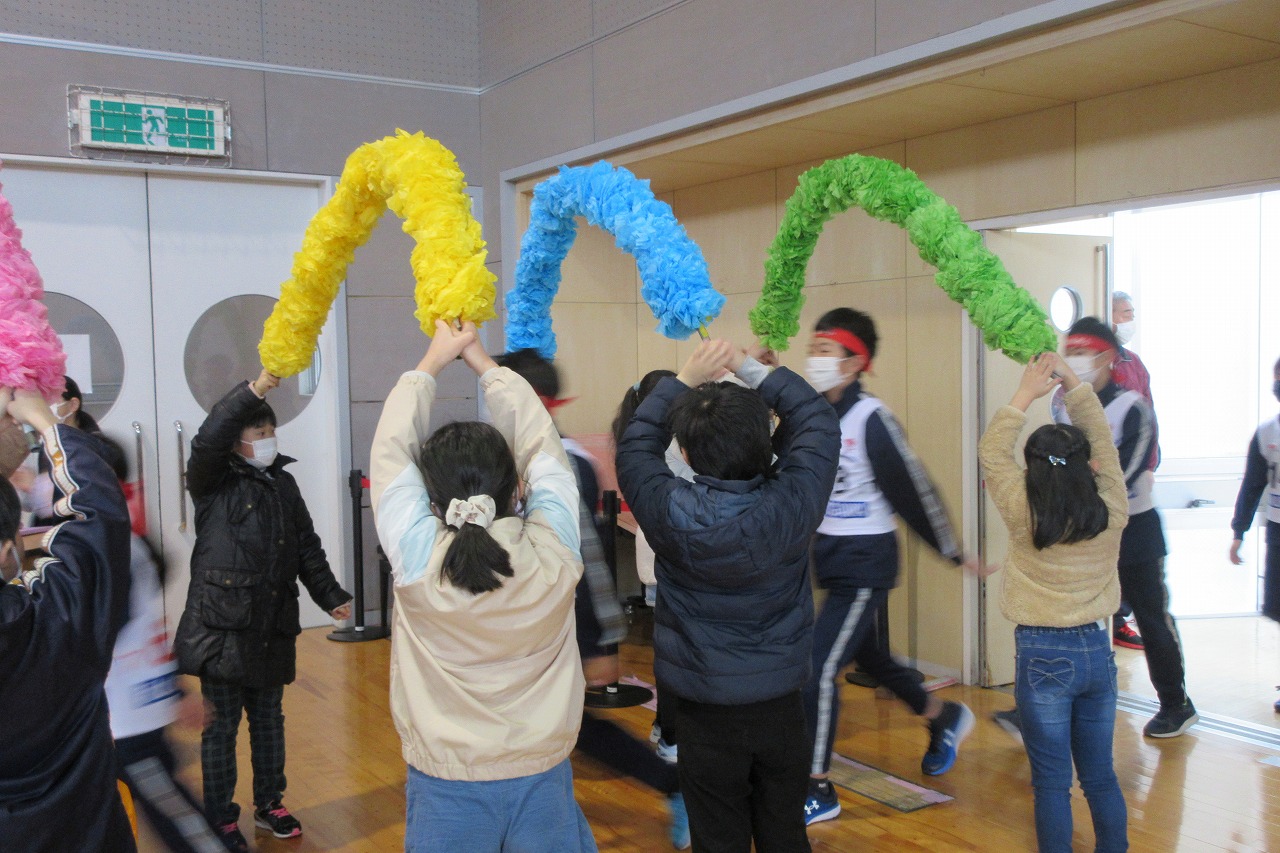 小学校の体育館で行われたスキー大会の壮行式にて、花紙を使って作られた複数のアーチを、各々2人一組で頭上に持ち上げる児童達と、その下を歩いてくるスキー大会参加者の児童達の様子を写した写真
