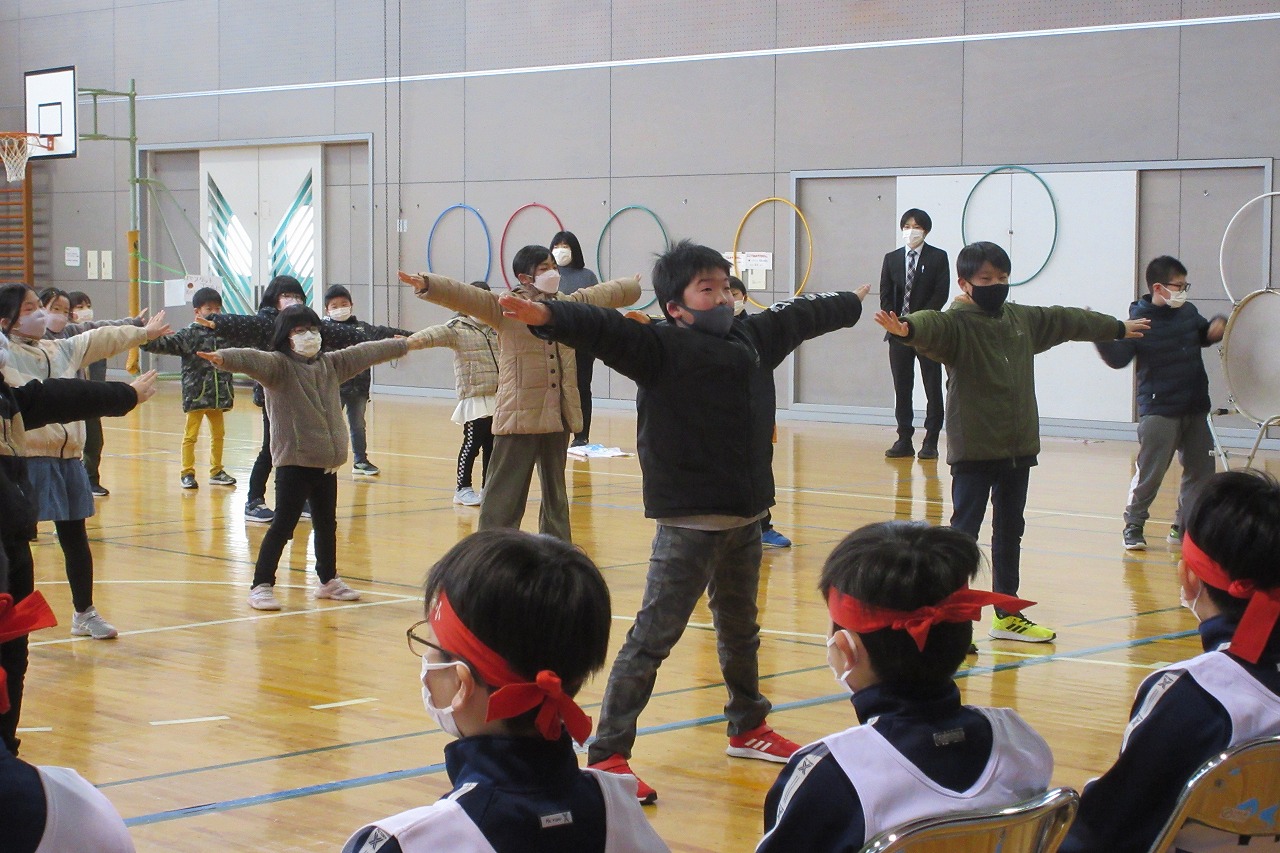 小学校の体育館で行われたスキー大会の壮行式にて、スキー大会に参加する児童達の前で、両手を広げてパフォーマンスを行う大勢の児童達の様子を写した写真