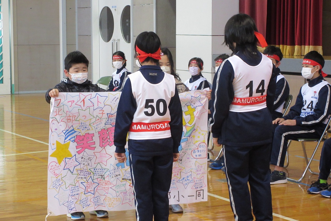 小学校の体育館で行われたスキー大会の壮行式にて、低学年の2人の児童から応援旗を渡される、スキー大会に参加する高学年の児童2人の様子を写した写真