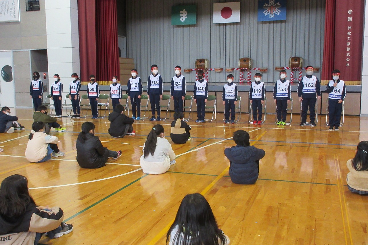 小学校の体育館で行われたスキー大会の壮行式にて、壇上の前に立つ、スキー大会に参加する14人の児童と、彼らに向かい合って、間隔を空けて床に並んで座っている大勢の児童達の様子を写した写真