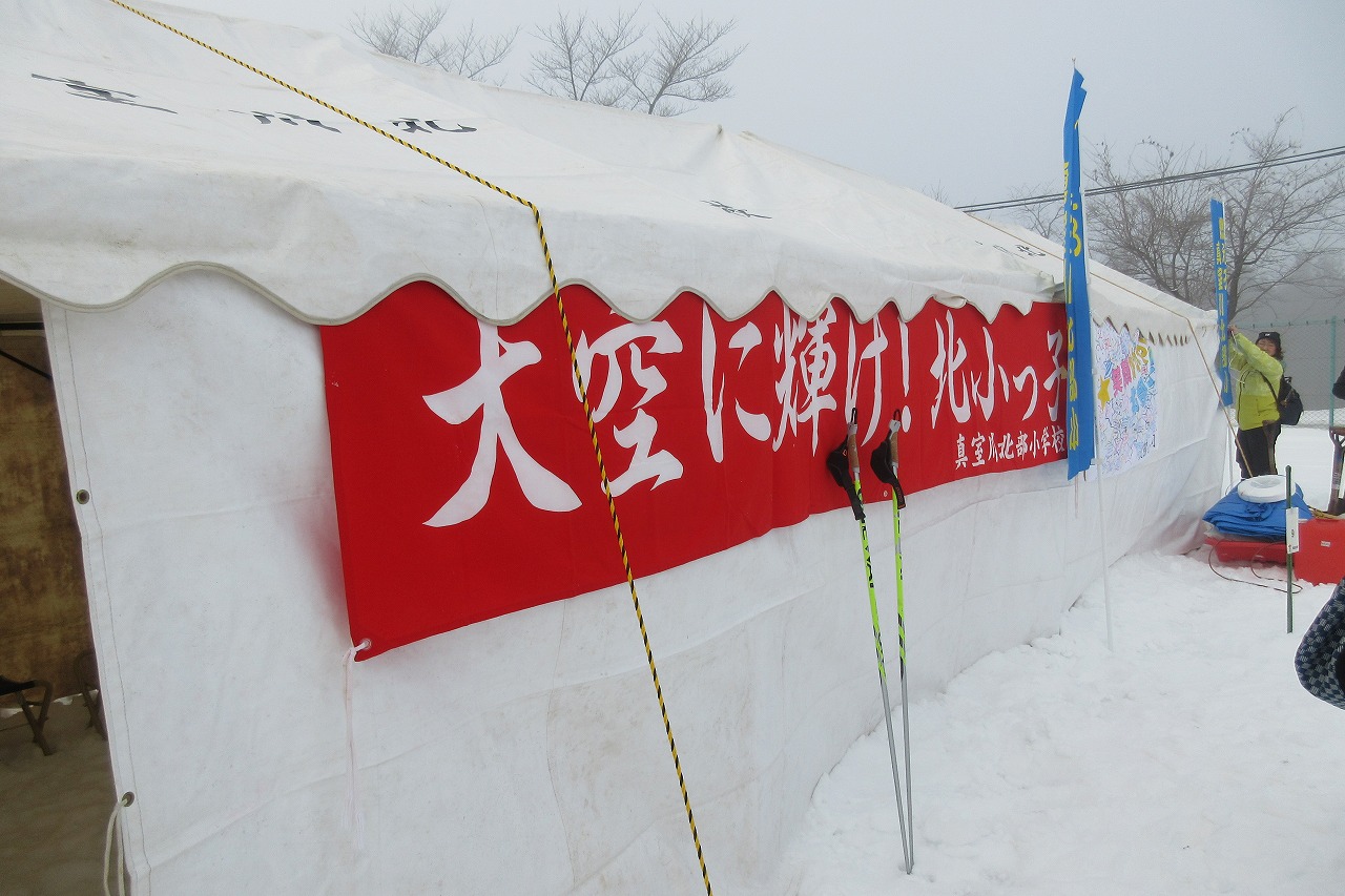 秋山スキー場で開催されたスキー大会にて、真室川北部小学校の待機場の白いテントに貼られた、「大空に輝け!北小っ子」と書かれた横長の赤い応援旗を写した写真