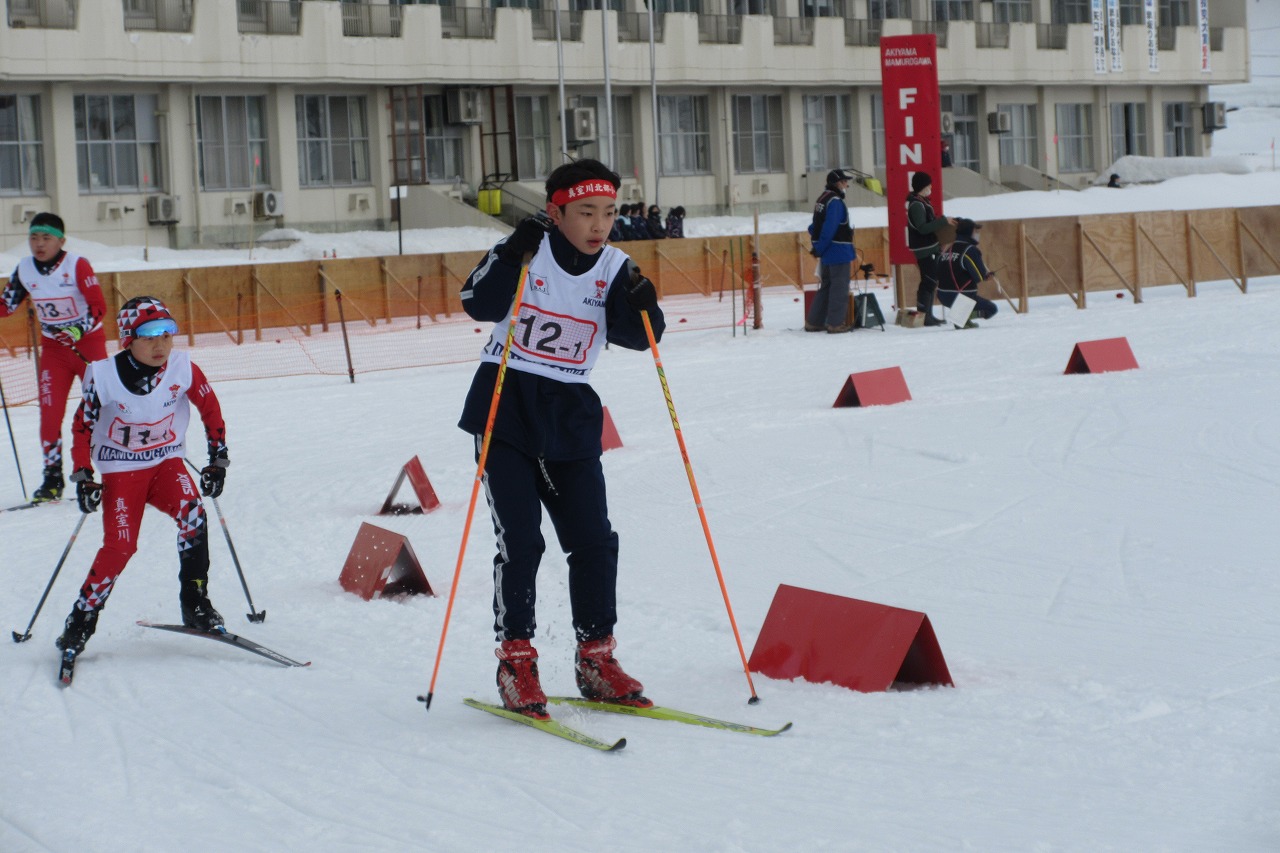 秋山スキー場で開催されたスキー大会にて、スキー板に乗って速さを競っている真室川北部小学校の男子児童と、他校の2人の男子児童の様子を写した写真