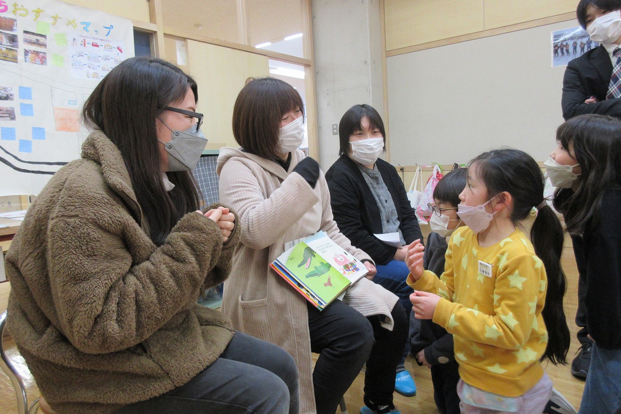 小学校の教室内で、椅子に座っているPTA研修部の3人の母親と、向かい合って膝立ちをしながら話をしている3人の児童の様子を写した写真