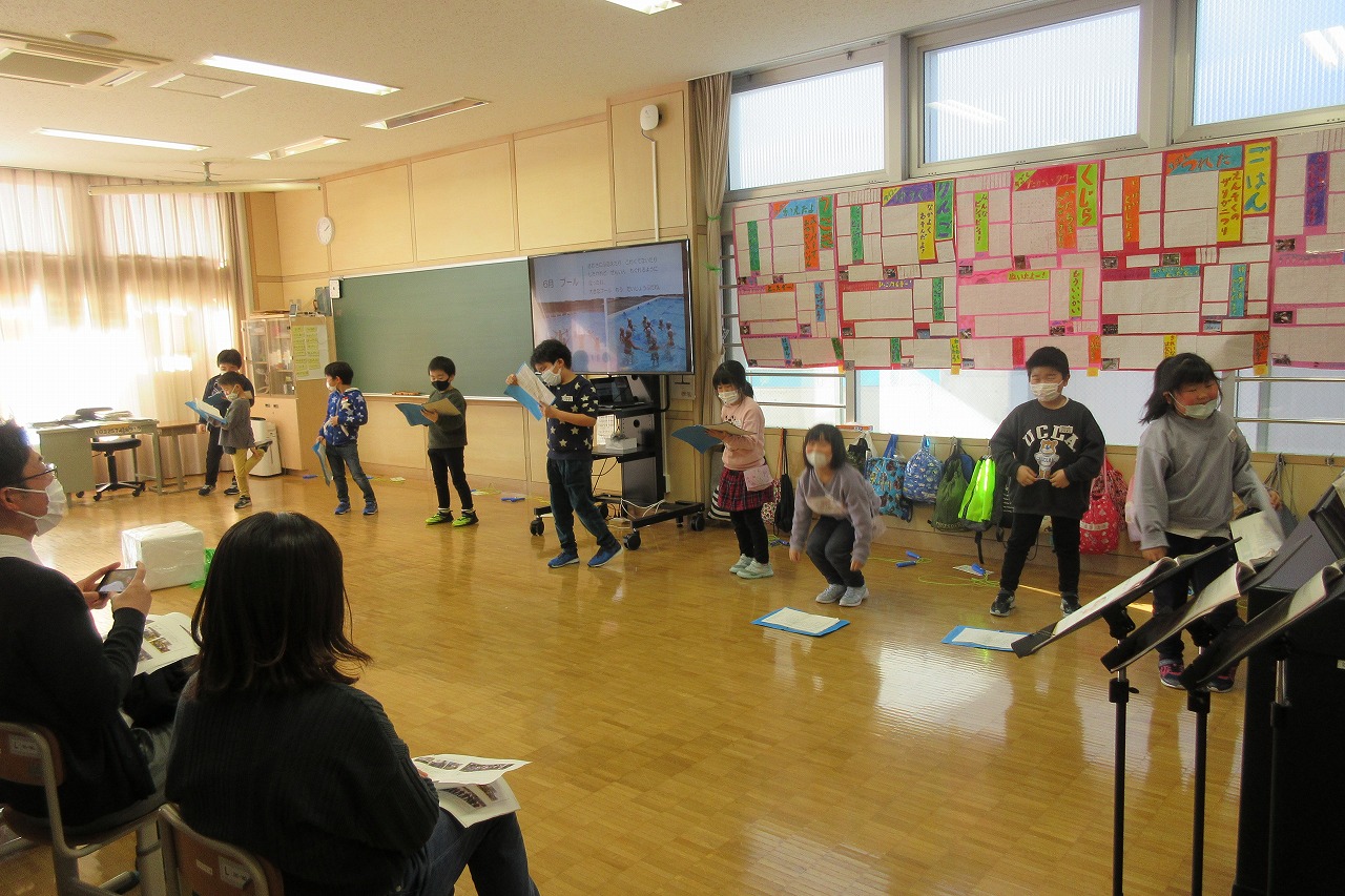 小学校の教室内で行われた授業参観にて、前に立って資料とモニターの写真を使いながら、一年の振り返りを行う9人の児童達の様子を写した写真